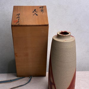 Może przedstawiać: Cylindryczny ceramiczny wazon o matowym, beżowym, nakrapianym wykończeniu z wyraźnymi czerwonobrązowymi wzorami przypominającymi płomienie. Obok stoi wysokie, prostokątne drewniane pudełko z czarną japońską kaligrafią i czerwoną pieczęcią.