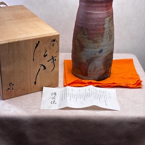 May include: A cylindrical ceramic vase with a reddish-brown and grey mottled glaze, accompanied by a wooden storage box featuring black calligraphy and a red seal, and a white paper document with the Japanese text 備前焼.