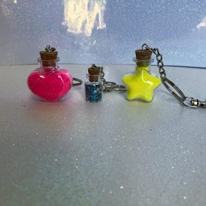 May include: Three glass bottle keychains with cork stoppers. One is a heart shape filled with neon pink glitter, one is a small cylinder filled with blue glitter, and one is a star shape filled with neon yellow glitter, each with a metal chain.