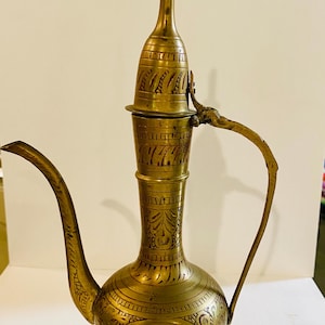 May include: A tall, metallic brass ewer featuring an elongated neck, a curved spout, and a hinged lid. The surface is decorated with intricate, etched floral and geometric patterns throughout the body, neck, and lid, with a sturdy base.