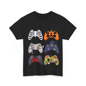 May include: A black short-sleeve t-shirt featuring a graphic print of six video game controllers styled as Halloween characters, including a skeleton ribcage, a jack-o'-lantern, a mummy, a witch, a vampire, and a green zombie face.