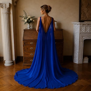 May include: A floor-length royal blue gown featuring a deep open back, wide shoulder straps, and a flowing, sheer chiffon cape-style overlay that extends into a full train, creating a dramatic silhouette with a solid, opaque bodice.