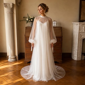 May include: A floor-length white dress featuring a sleeveless bodice with a sweetheart neckline, layered under a sheer tulle overlay. The overlay includes a high illusion neckline and long, voluminous bishop sleeves with gathered cuffs.