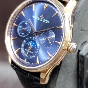 Könnte beinhalten: Eine roségoldene Jaeger-LeCoultre Armbanduhr mit tiefblauem Sonnenschliff-Zifferblatt, goldenen Stundenmarkierungen, drei Hilfszifferblättern, Mondphasenanzeige und schwarzem Lederarmband. Das Zifferblatt zeigt den Schriftzug Jaeger-LeCoultre und Automatique.