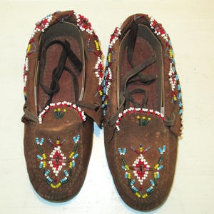 May include: A pair of brown leather moccasins with intricate beadwork in red, white, blue, and yellow. The moccasins have a geometric pattern and are tied with leather laces.