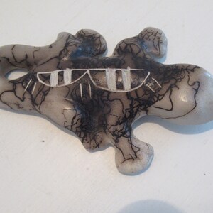 May include: A ceramic lizard figurine with black and white painted details. The lizard is in a light gray color with black lines and a white stripe across its back.