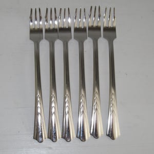 May include: Six silver forks with ornate handles. The forks are arranged in a row, with the handles pointing towards the viewer.