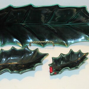 May include: A set of three ceramic serving dishes in the shape of holly leaves. The largest is a long tray, and the other two are smaller dishes. All are dark green with red berry accents.
