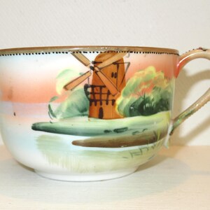 May include: A white teacup with a hand-painted landscape scene featuring a windmill, trees, and a sunset. The cup has a decorative handle and a brown rim with small dotted accents. The cup is a vintage item.