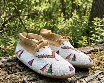 May include: A pair of tan suede moccasins featuring white seed beadwork across the vamps and sides. The beadwork displays geometric patterns in red, green, orange, and blue. Each shoe includes a tan suede lace tied at the center front.