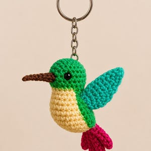 Puede incluir: Un llavero de colibrí hecho a mano con ganchillo. El pájaro tiene una cabeza verde, un ala turquesa, un vientre amarillo y una cola rosa. Tiene un pico marrón y un ojo negro. El llavero incluye un anillo y una cadena de metal.
