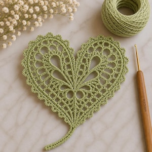 Può includere: Un centrino all'uncinetto a forma di cuore color verde salvia con un intricato motivo in pizzo e bordi smerlati. Accanto al centrino si trovano un gomitolo di filo verde abbinato e un uncinetto in legno con la misura 0,60 mm incisa.