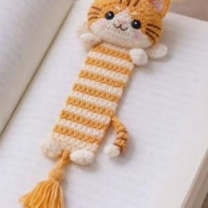 May include: A crocheted bookmark shaped like a ginger tabby cat with a striped body, white paws, and a small tail. The cat head features embroidered black eyes, a pink nose, and whiskers. A yellow yarn tassel is attached to the bottom end.