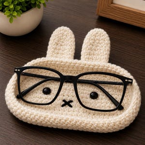 May include: A cream-colored crochet tray shaped like a rabbit head with two upright ears, two black circular eyes, and a black stitched X-shaped mouth. A pair of black-framed eyeglasses rests horizontally across the center of the crochet tray.