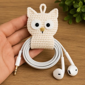 May include: A cream-colored crocheted owl cable organizer featuring a small loop at the top, two large white eyes with black pupils, and a small yellow beak. The owl is wrapped around a coiled white wired earphone set with a standard audio jack.
