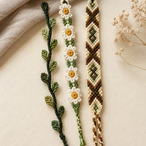 Op de afbeelding: Drie handgemaakte macramé armbanden met verschillende ontwerpen. De eerste is een donkergroene wijnstok met kleine blaadjes. De tweede toont een rij witte madeliefjes met gele harten. De derde heeft een geometrisch patroon in crème, bruin en groen.