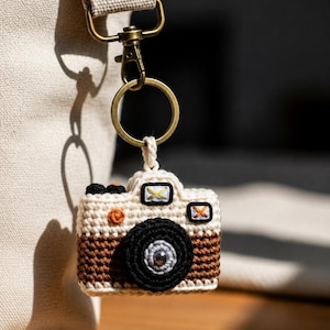 Pode incluir: Um porta-chaves de câmara de croché em creme, castanho e preto, com uma lente prateada. O porta-chaves está preso a um anel dourado e a uma alça de saco de lona. A câmara tem detalhes bordados e um círculo laranja decorativo.