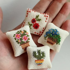 May include: Four miniature cross-stitch pillows, each featuring a different floral design. The pillows are square, with a cream-colored background and colorful embroidered flowers, including roses, petunias, and cacti. The pillows are held in a hand.