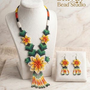 Può includere: Un set di collana e orecchini in perline con un grande ciondolo centrale a forma di fiore giallo e arancione, dettagli verdi e frange pendenti. La collana presenta motivi floreali e geometrici. Il testo riporta Shivay Bead Studio.