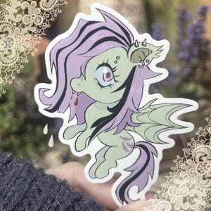 May include: A die-cut sticker featuring a pale green, winged creature with long, flowing lavender and black hair. The character has a single large eye with a spiral pupil, small fangs with red droplets, and a bat-like wing. It features purple bat-shaped markings on its flank and a spiked ear accessory.