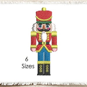 Può includere: Un disegno ricamato di uno schiaccianoci con giacca e cappello rossi, pantaloni blu e stivali neri con dettagli dorati. La figura ha barba e baffi bianchi. Il testo 6 Sizes è stampato in nero a sinistra della figura dello schiaccianoci.