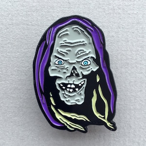May include: A purple and black enamel pin featuring a cartoon character with a skull-like face, a wide grin, and glowing blue eyes.