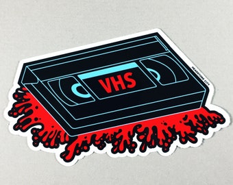 May include: A black VHS tape sticker with the word "VHS" in red. The tape is outlined in light blue and has a red splatter design beneath it. The sticker has the text "HEADSTONE CITY" in the upper right corner.