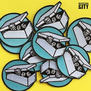 May include: A set of eight embroidered iron-on patches featuring a white box with a black outline, a black lid, and a black and white design inside. The box is open and the lid is propped up. The patches are on a yellow background. The text "HEADSTONE CITY" is in black letters on a white background in the upper right corner of the image.