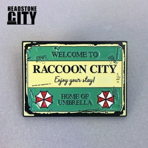 May include: A rectangular metal sign with a black border. The sign has a green top and bottom section with the words "WELCOME TO" and "HOME OF UMBRELLA". The center section is yellow with "RACCOON CITY" and "Enjoy your stay!" written in black. Two red and white umbrella logos are present.