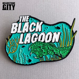 May include: A teal and black enamel pin featuring the text "THE BLACK LAGOON" in white, with a green creature design. The pin includes yellow and green aquatic plant illustrations and the text "HEADSTONE CITY" at the top.