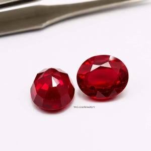 Puede incluir: Dos piedras preciosas rojas vibrantes facetadas con un corte brillante. Una piedra es redonda y la otra es de forma ovalada. El texto VeloraGemsArt está impreso en letras negras pequeñas entre las dos piedras sobre un fondo blanco liso.