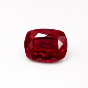 May include: A vibrant, deep red gemstone with a cushion cut, featuring multiple triangular and trapezoidal facets that create a brilliant, reflective surface. The stone has a rectangular shape with rounded corners and a rich, saturated colour.