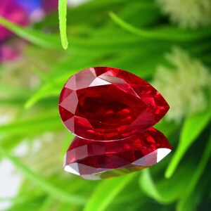 Peut inclure: Une pierre précieuse rouge taillée en forme de poire avec des facettes brillantes, présentant des teintes cramoisies profondes et des angles géométriques nets. La pierre est positionnée pour montrer sa surface réfléchissante et transparente.