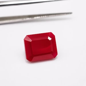 May include: A vibrant red gemstone cut into a rectangular emerald shape with faceted edges. The stone displays a deep, saturated crimson hue and sits positioned below a pair of metallic silver tweezers, showcasing its polished, reflective surface.
