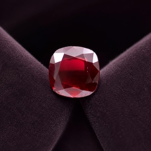 Pode incluir: Uma pedra preciosa de rubi com lapidação almofada, cor vermelha profunda e superfície facetada. A pedra exibe reflexos de luz internos e uma forma simétrica, apresentando um acabamento polido que destaca o seu tom vermelho vibrante e saturado.
