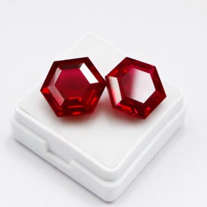 May include: Two vibrant red gemstones cut into a hexagonal shape with faceted surfaces. The stones are displayed side by side on a small, square white plastic base, showcasing their deep crimson colour and geometric symmetry in clear detail.