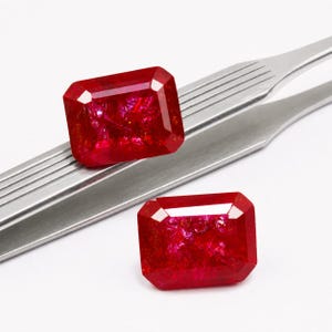 Può includere: Due gemme rosso intenso con taglio a raggiera. Le gemme sono esposte su una pinzetta d'argento su sfondo bianco. Le gemme hanno una forma rettangolare con bordi sfaccettati.