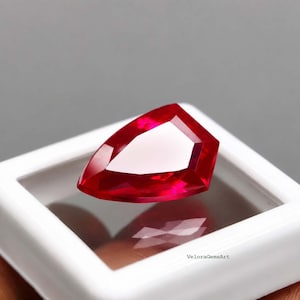 Puede incluir: Una gema de color rojo vibrante con un corte facetado en forma de pera, que muestra bordes geométricos afilados y una superficie pulida. La gema está colocada dentro de una caja expositora blanca cuadrada con el texto VeloraGemsArt en la base.