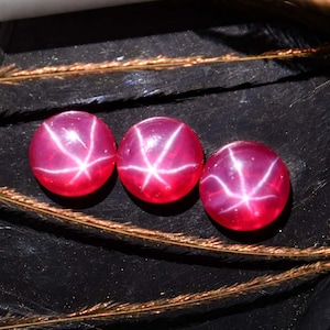 Puede incluir: Tres rubíes redondos tallados en cabujón con efecto estrella. Las gemas son de color rojo intenso con un patrón de estrella de seis rayos. Las gemas se exhiben sobre una superficie oscura con hierba seca.