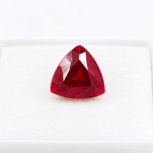 Peut inclure: Une pierre précieuse d'un rouge profond et vibrant, taillée en forme triangulaire avec de multiples facettes qui reflètent la lumière. La pierre présente une teinte cramoisie saturée et brillante ainsi qu'une silhouette géométrique nette.