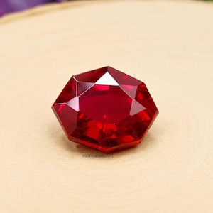 Könnte beinhalten: Ein leuchtend roter Edelstein mit einem facettierten, sechseckigen Schliff, der das Licht auf seiner polierten Oberfläche reflektiert. Der Stein hat scharfe, geometrische Kanten und einen tiefen Karmesinton, mit dem Text VeloraGemsArt in kleiner Schrift.