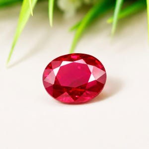 Puede incluir: Una gema roja de corte ovalado con una superficie facetada que refleja la luz. La piedra muestra un tono carmesí profundo y saturado, posicionada sobre un fondo neutro con follaje verde visible en la parte superior del encuadre.