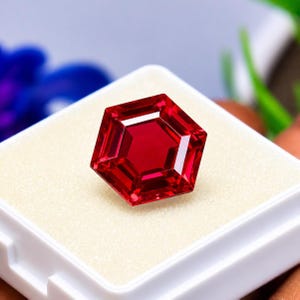 Puede incluir: Una gema de color rojo intenso y vibrante tallada en una forma hexagonal precisa con múltiples facetas que reflejan la luz. La gema se muestra descansando sobre una base de espuma de color crema dentro de un pequeño estuche cuadrado blanco.
