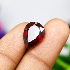 Può includere: Una gemma rosso scuro a forma di pera con una superficie sfaccettata che riflette la luce. La pietra presenta una parte superiore appuntita e una base arrotondata, mostrando un colore cremisi ricco e saturo in tutta la sua struttura cristallina.