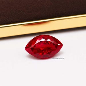 May include: A marquise-cut gemstone with a vibrant, deep red color and a faceted surface that reflects light. The stone is positioned on a white surface with the text VeloraGemsArt printed in small black letters near the bottom right edge.