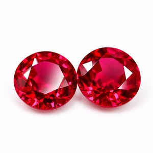 Könnte beinhalten: Zwei rund geschliffene, leuchtend rote Edelsteine mit einer facettierten Oberfläche, die das Licht reflektiert. Diese kreisförmigen Steine weisen einen tiefen, gesättigten karmesinroten Farbton und eine polierte Oberfläche mit klarem Muster auf.