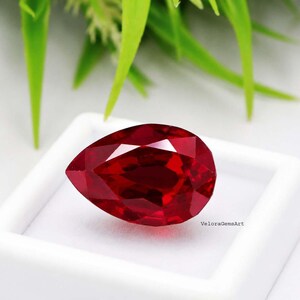 May include: A pear-shaped, faceted red gemstone with a deep crimson hue and a brilliant, reflective surface. The stone is positioned inside a square white display box, with the text VeloraGemsArt printed in small black letters on the base.
