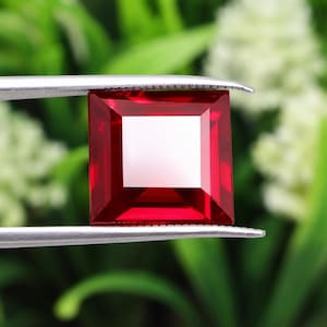Può includere: Una pietra preziosa di colore rosso vibrante a taglio quadrato tenuta da una pinzetta in metallo argentato. La pietra presenta una superficie sfaccettata con bordi netti e un aspetto brillante e trasparente che riflette la luce sulle sue facce.