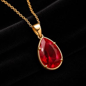 Puede incluir: Un collar de oro con un colgante de piedra preciosa roja tallada en forma de pera, sostenido por un engaste de oro de cuatro garras. El colgante cuelga de una delicada cadena de oro, mostrando un color rojo vibrante y un acabado dorado pulido.
