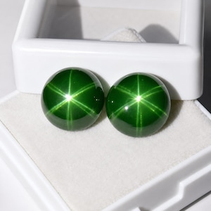 Könnte beinhalten: Zwei runde, im Cabochon-Schliff gearbeitete grüne Edelsteine mit einem ausgeprägten sechsstrahligen Sterneffekt, bekannt als Asterismus. Die Steine sind glatt, poliert und zeigen eine tiefgrüne Farbe mit hellen, weißen Lichtreflexen.